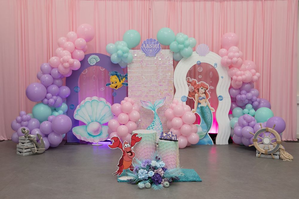 Little Mermaid birthday party backdrop with pink, purple, and teal balloon arch, Ariel cutout, and seashell decorations.