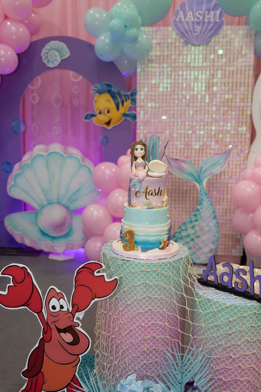 Little Mermaid themed birthday cake with mermaid topper named Aashi, surrounded by balloon arch and Sebastian decoration.