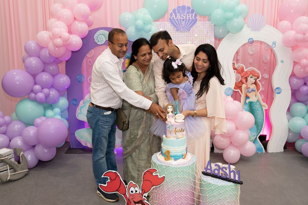 Family celebrating a Little Mermaid themed birthday party with balloon arch and Ariel decorations while cutting cake.