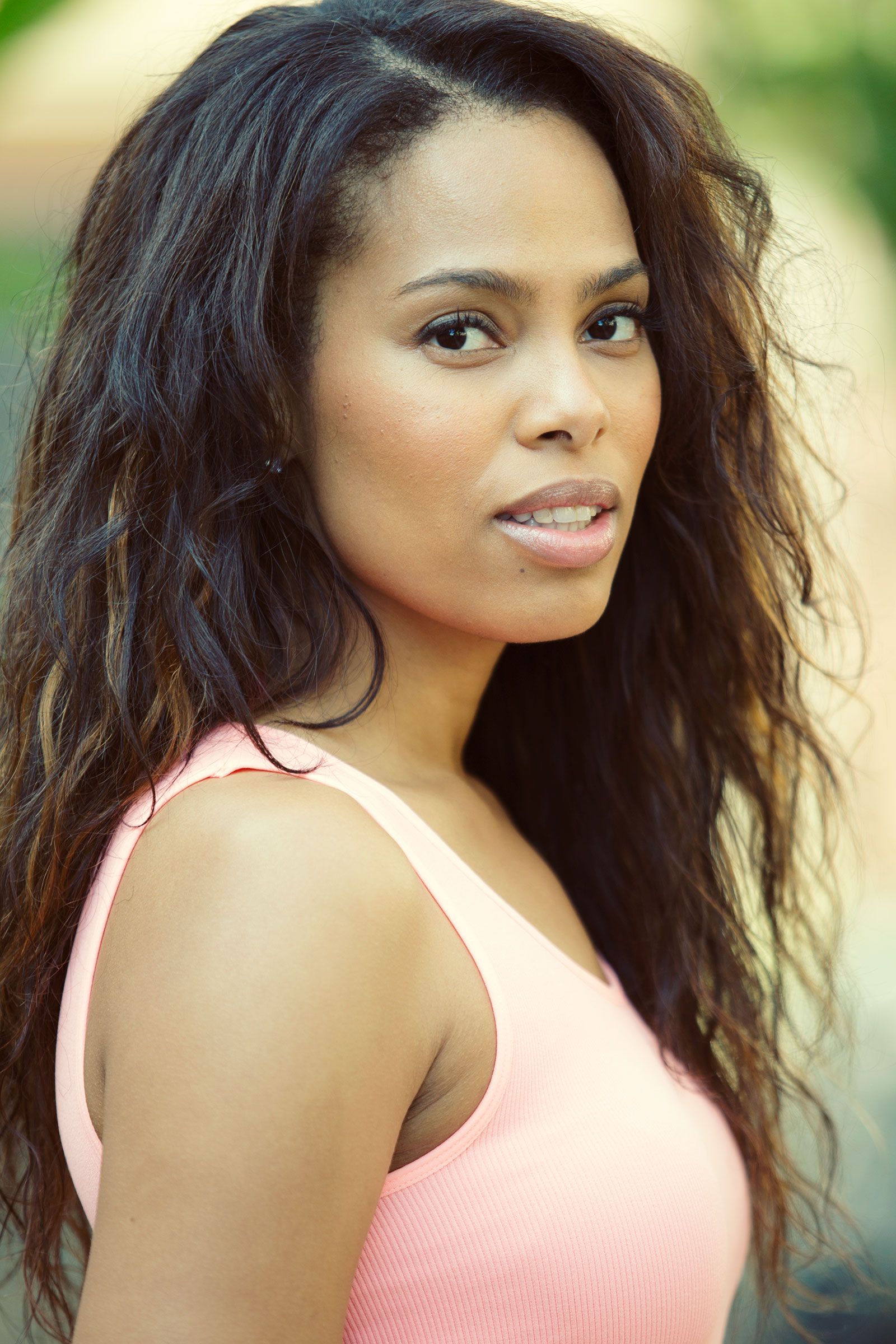Woman with long wavy dark hair in pink tank top smiling outdoors.