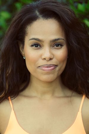 Close-up portrait of a woman with dark wavy hair and subtle smile wearing an orange top outdoors.