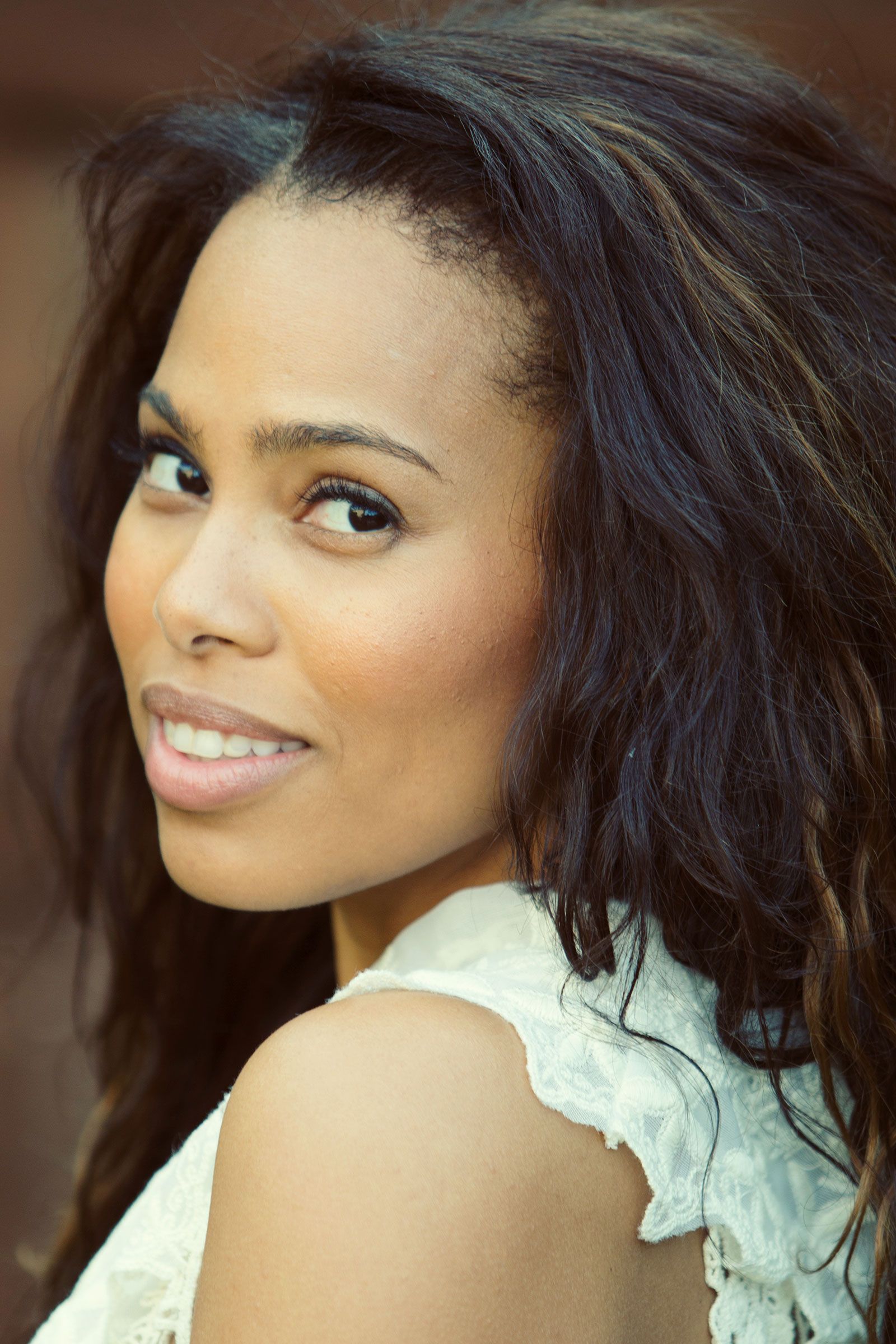 Woman with tousled dark hair smiling over shoulder wearing white lace top in warm outdoor setting.