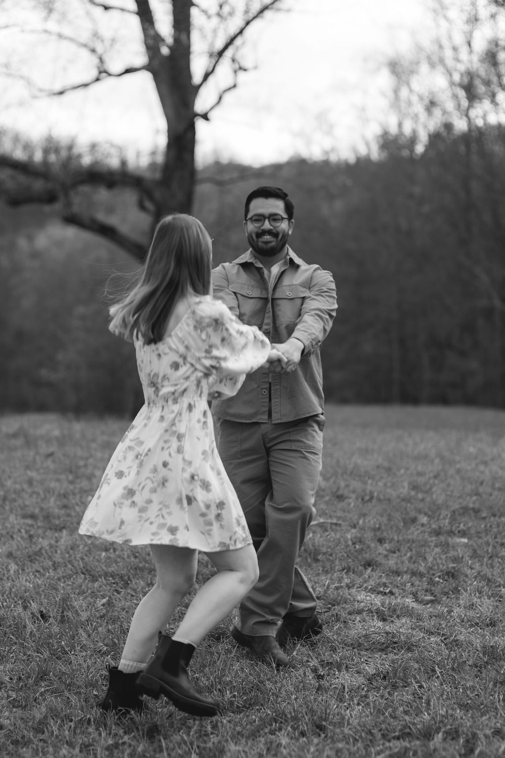 Black and white photo of a couple dancing and holding hands in a grassy field with trees in the background.