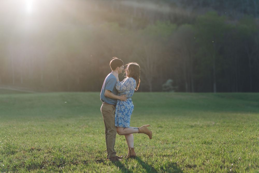 Couple sharing a romantic kiss in a sunlit green meadow, surrounded by trees, with golden backlight creating a dreamy atmosphere.