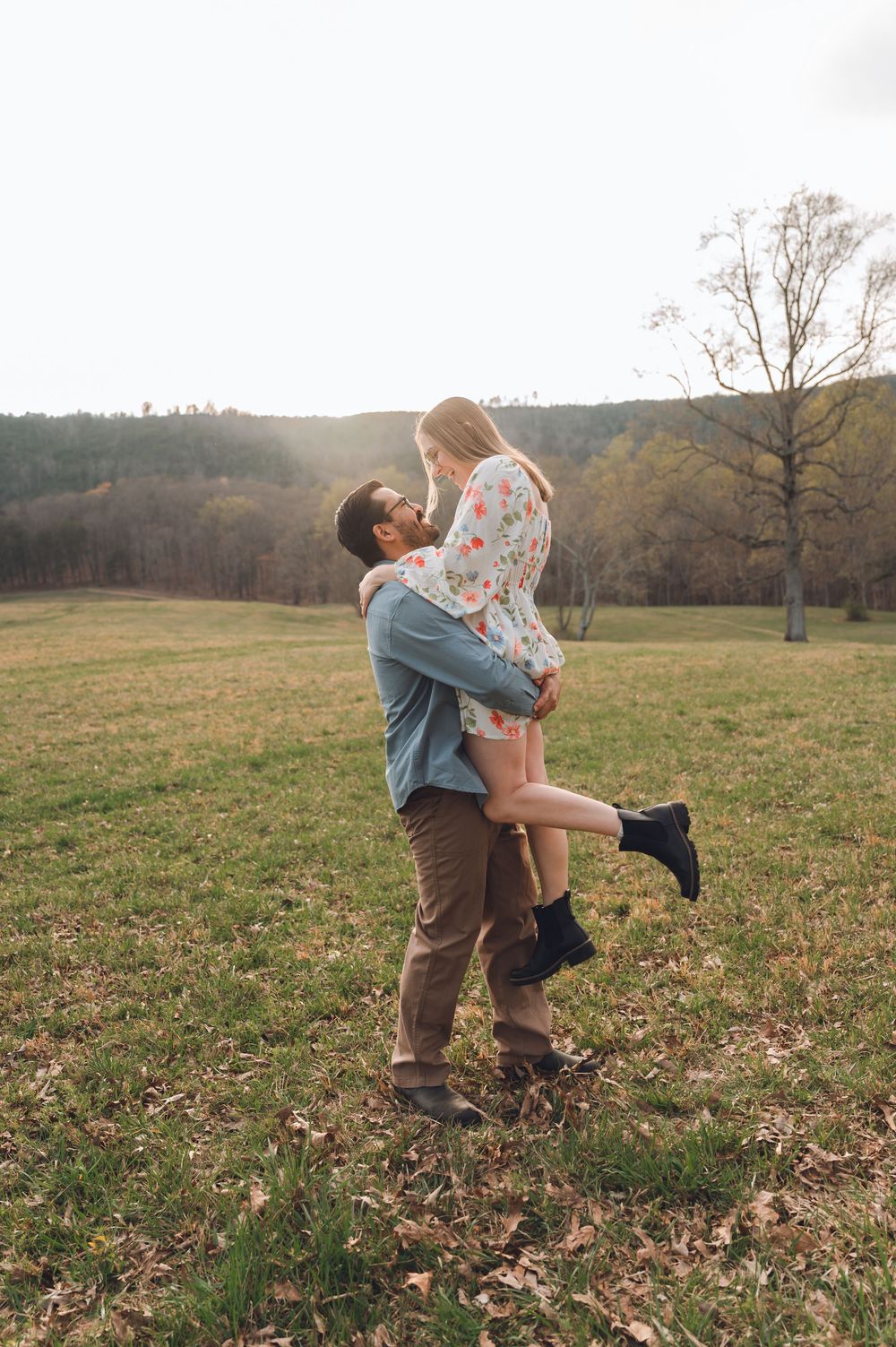A man lifts a woman in a floral dress in a scenic green field, sharing a romantic moment outdoors.
