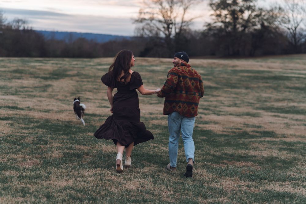 A couple walks hand-in-hand across a grassy field at dusk with a dog beside them, viewed from behind.