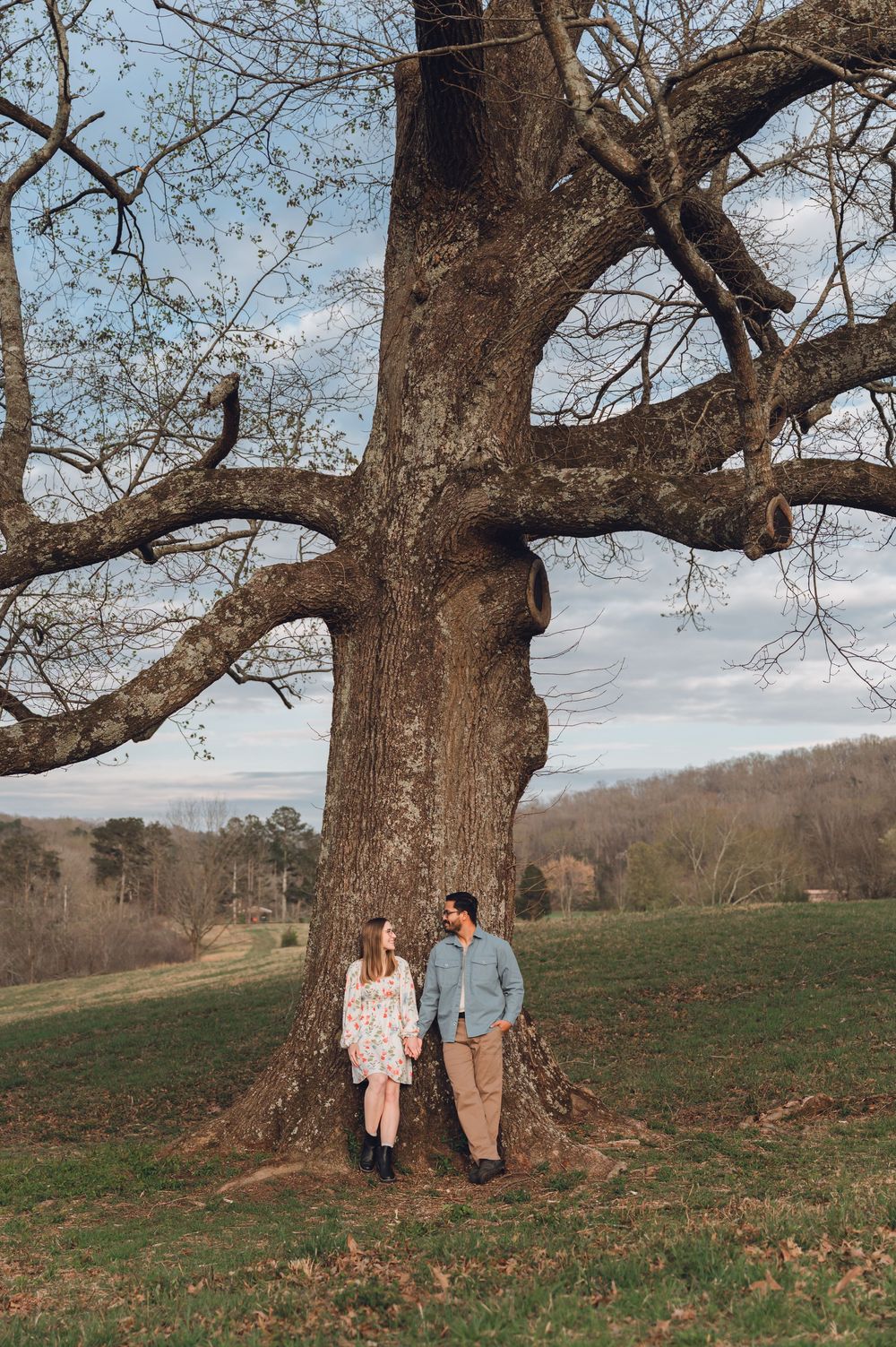 Couple holding hands leaning against a large tree in a green field with rolling hills and cloudy sky backdrop.