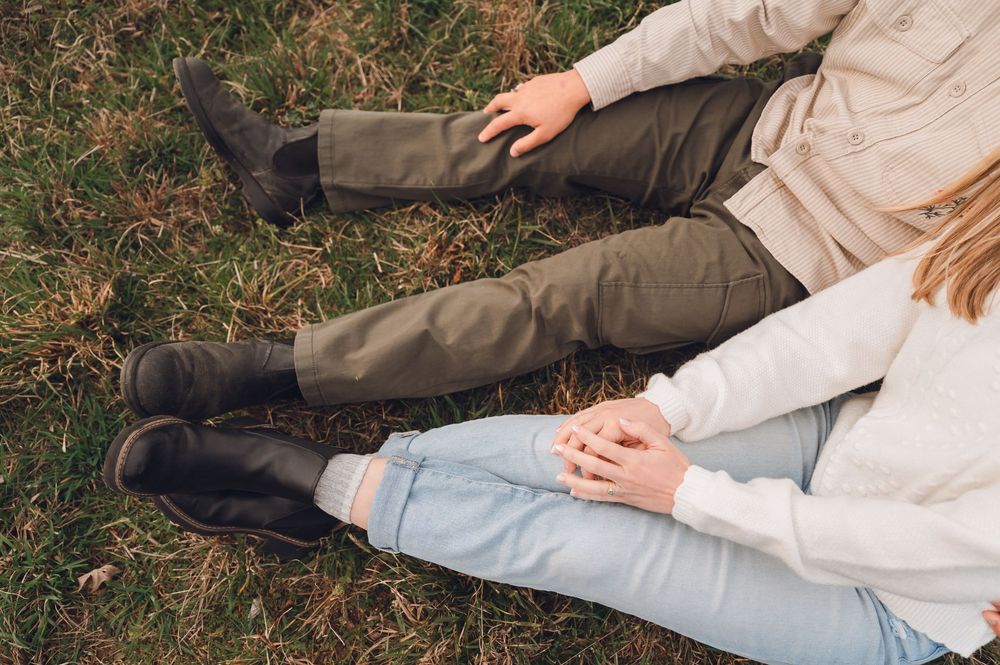 Couple sitting on grass holding hands, wearing dark boots, olive pants and light jeans, outdoor fall setting.