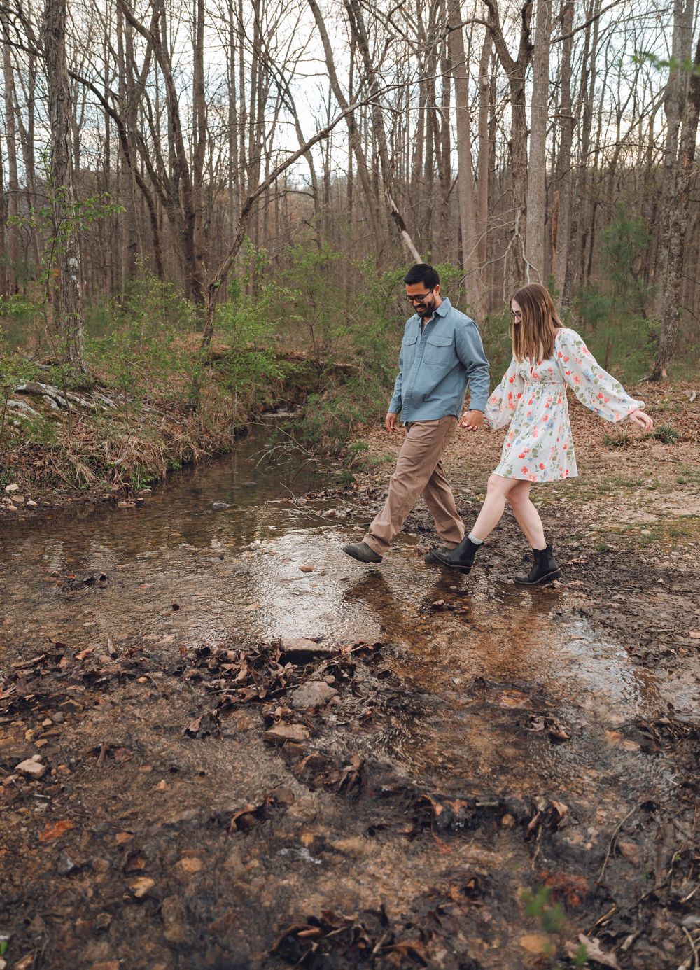 A couple holds hands while crossing a creek in a wooded forest during early spring.