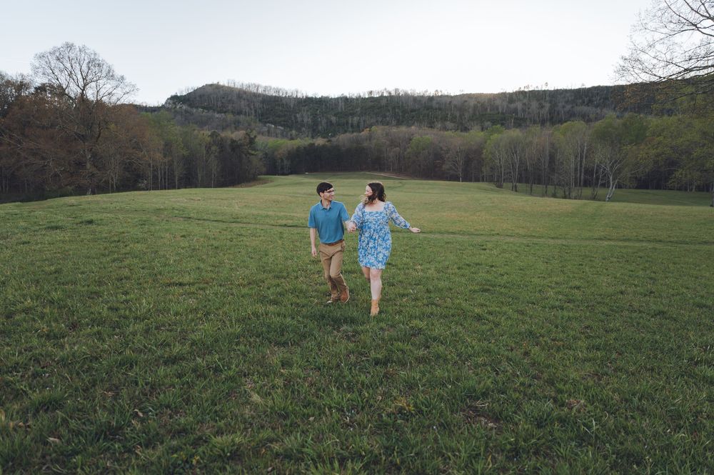 Couple holding hands running through a lush green meadow with rolling hills and trees in the background.
