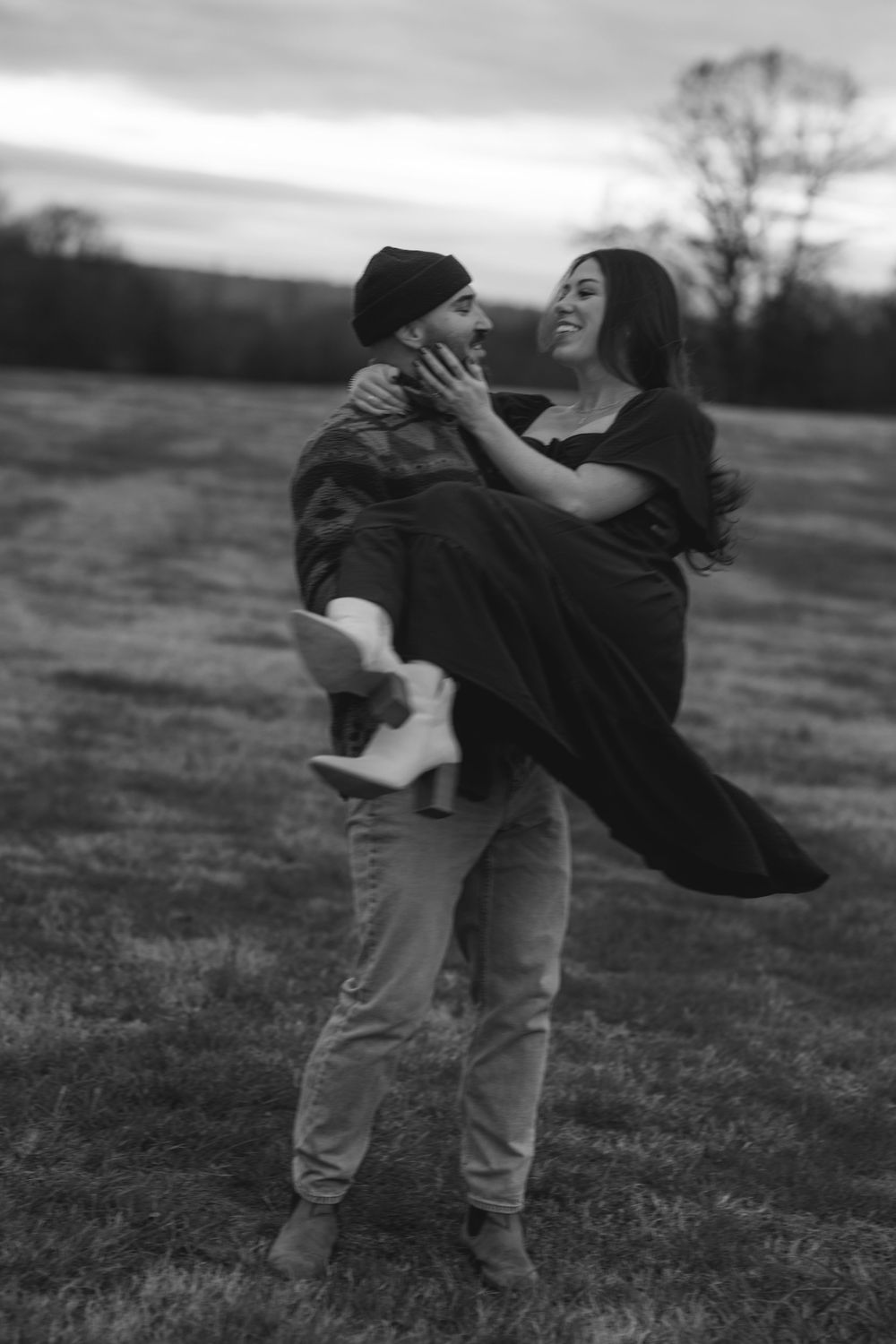 Black and white photo of a man carrying a woman in a field at dusk, both smiling and sharing a romantic moment.