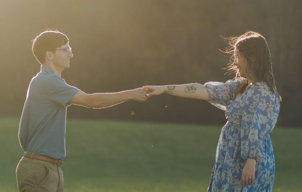 Couple holding hands at golden hour in a sunlit green field, woman in floral dress, man in casual attire.