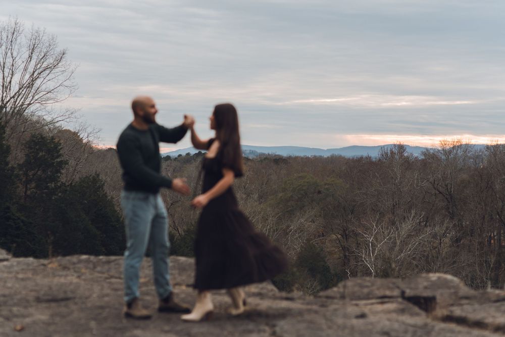 Couple dancing on rocky overlook at dusk with scenic mountain and forest views in the background.
