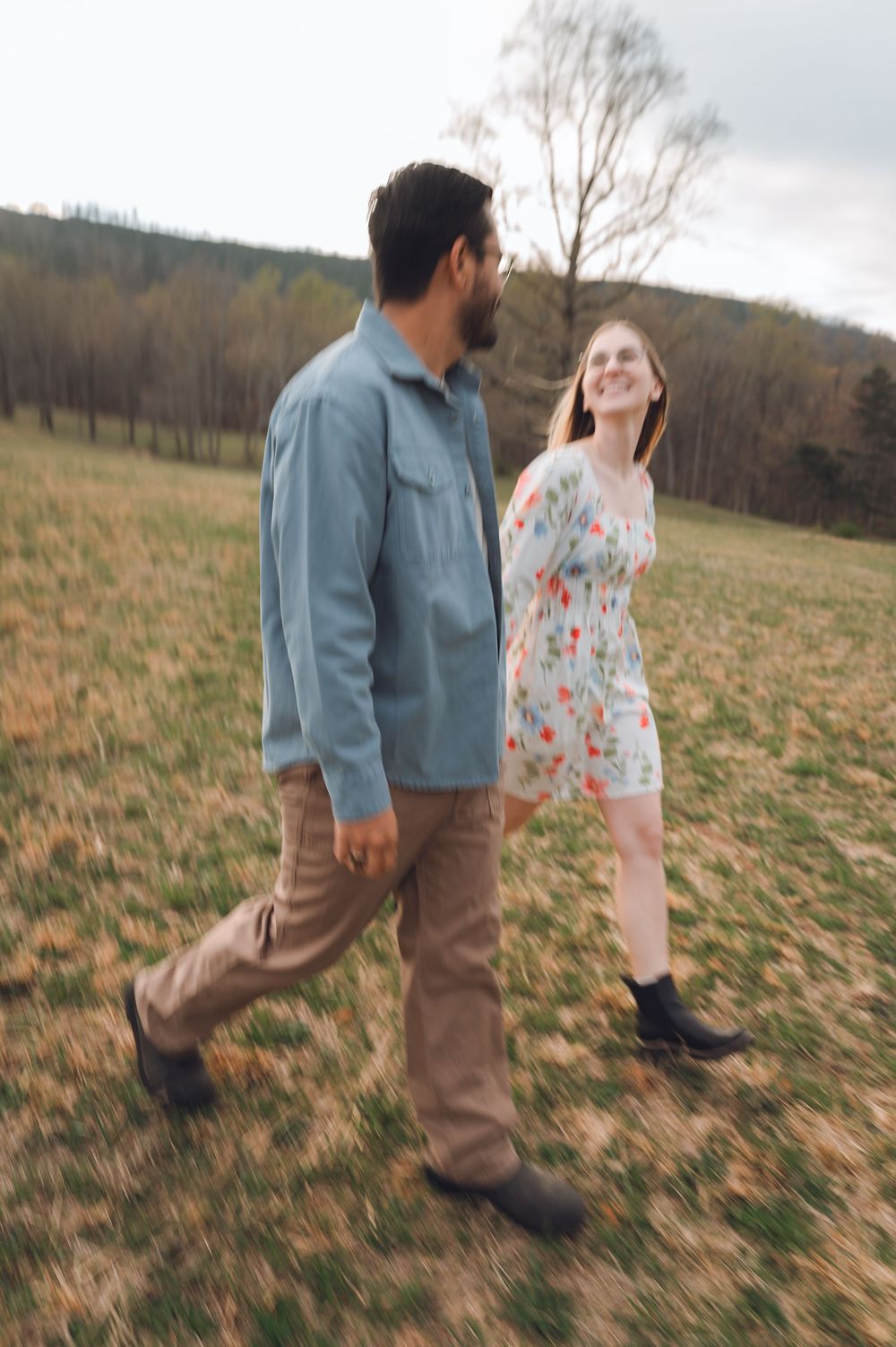 A couple walks hand-in-hand through a grassy field, woman in floral dress smiling at bearded man in blue shirt.