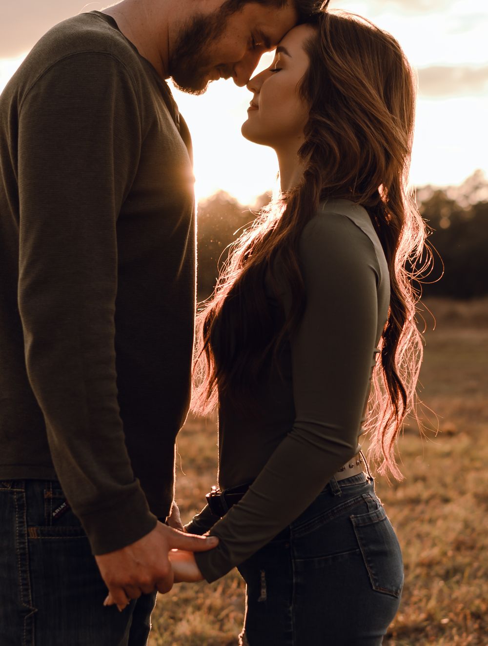 Couple holding hands and touching foreheads at golden sunset in a field, romantic outdoor portrait photography.