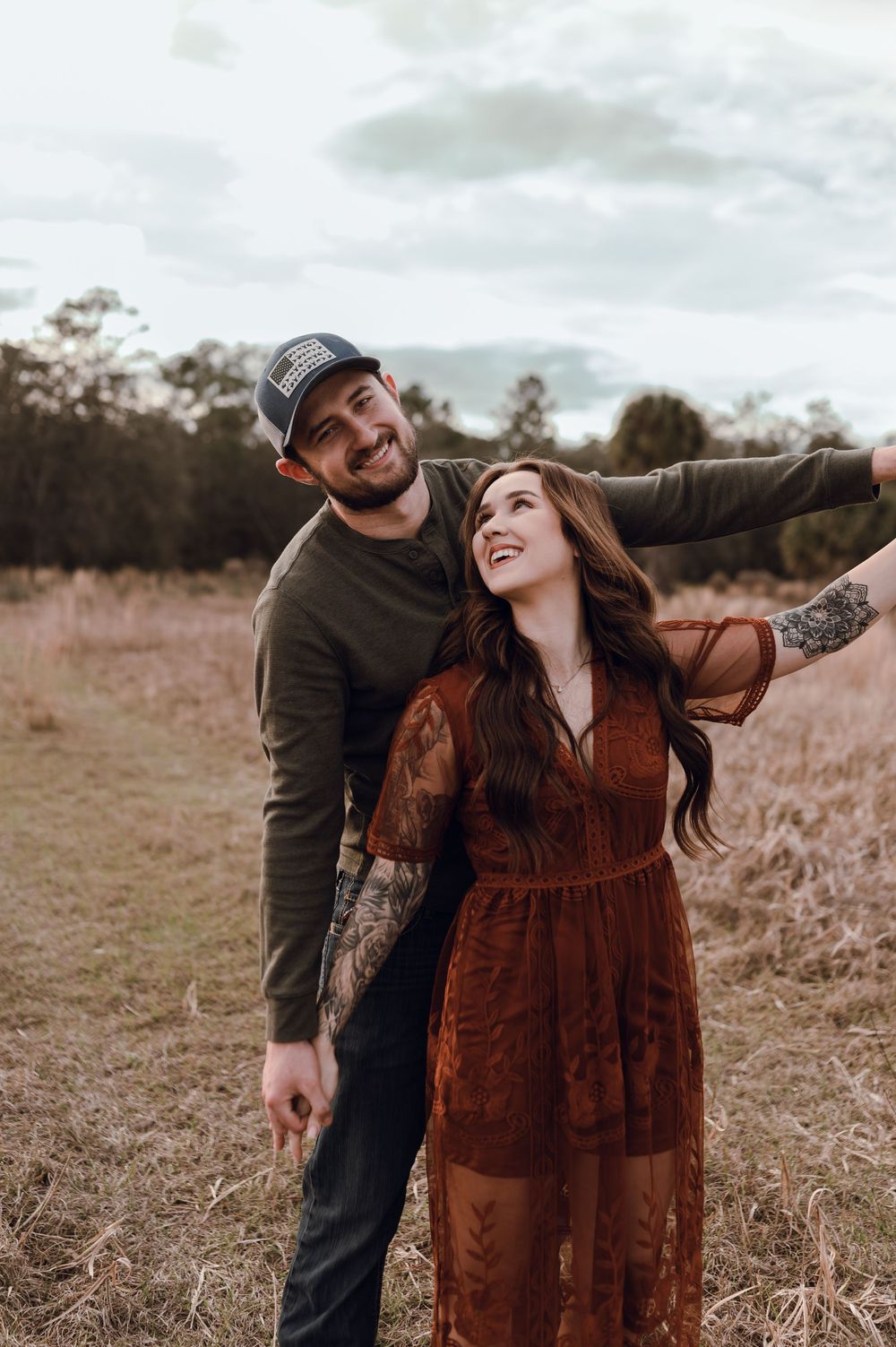 A smiling couple poses in a rustic field, woman in rust lace dress, man in green sweater and cap, arms outstretched.