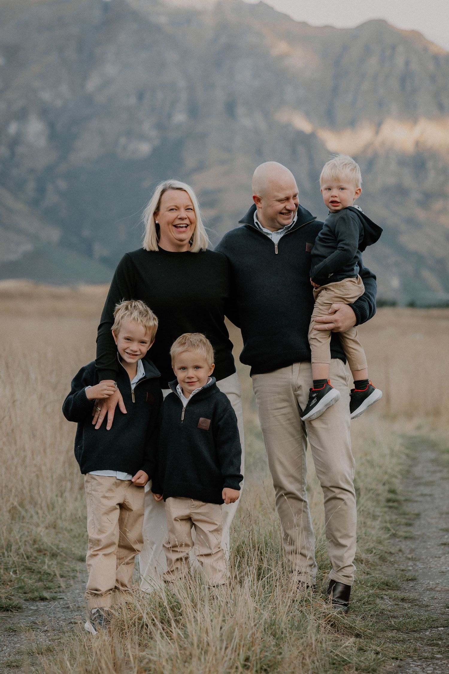 Happy family of five posing outdoors in matching black and tan outfits with mountain scenery in background.