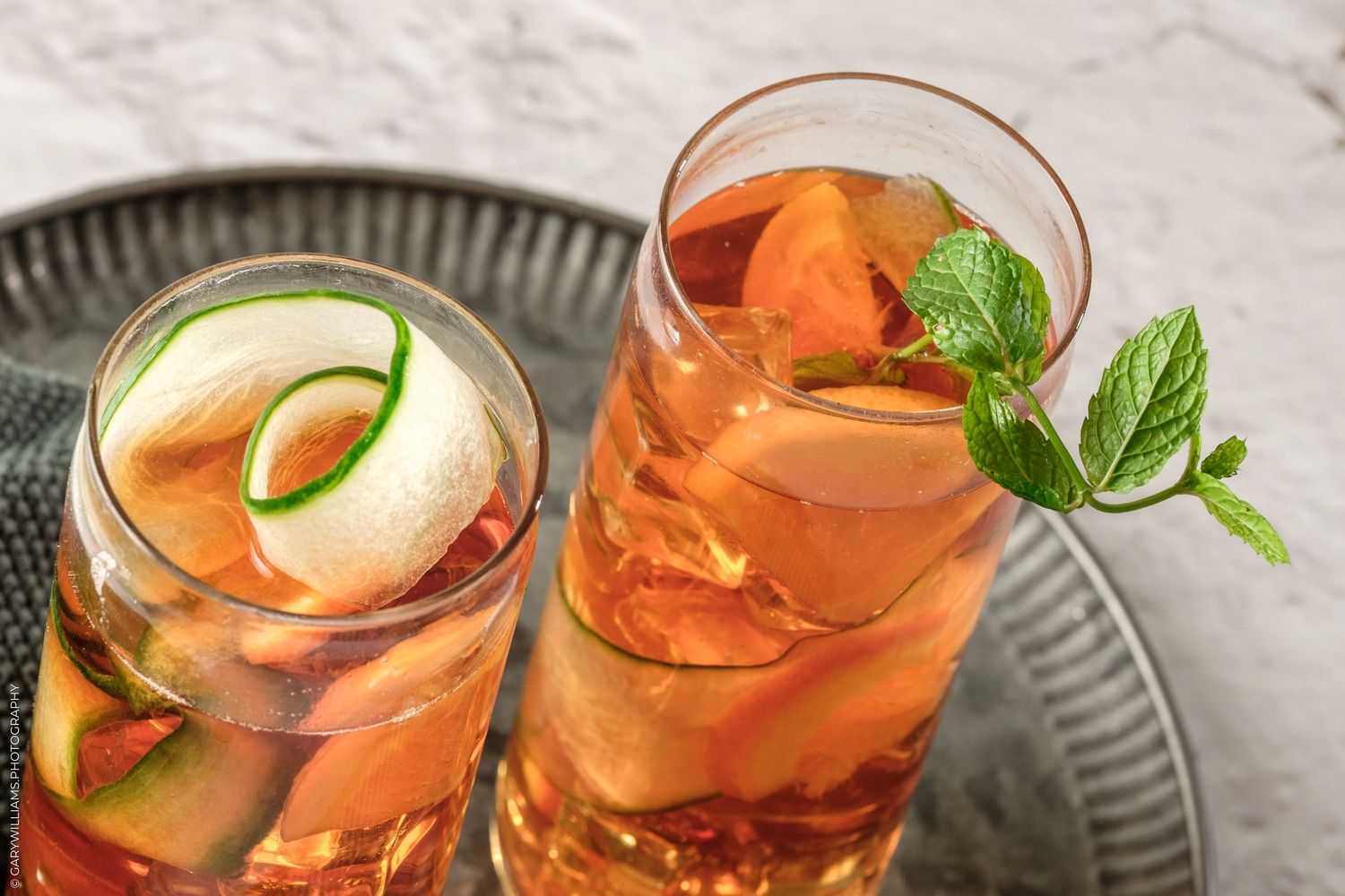 Two tall glasses of iced peach tea cocktails garnished with fresh mint leaves and fruit slices on a silver tray.