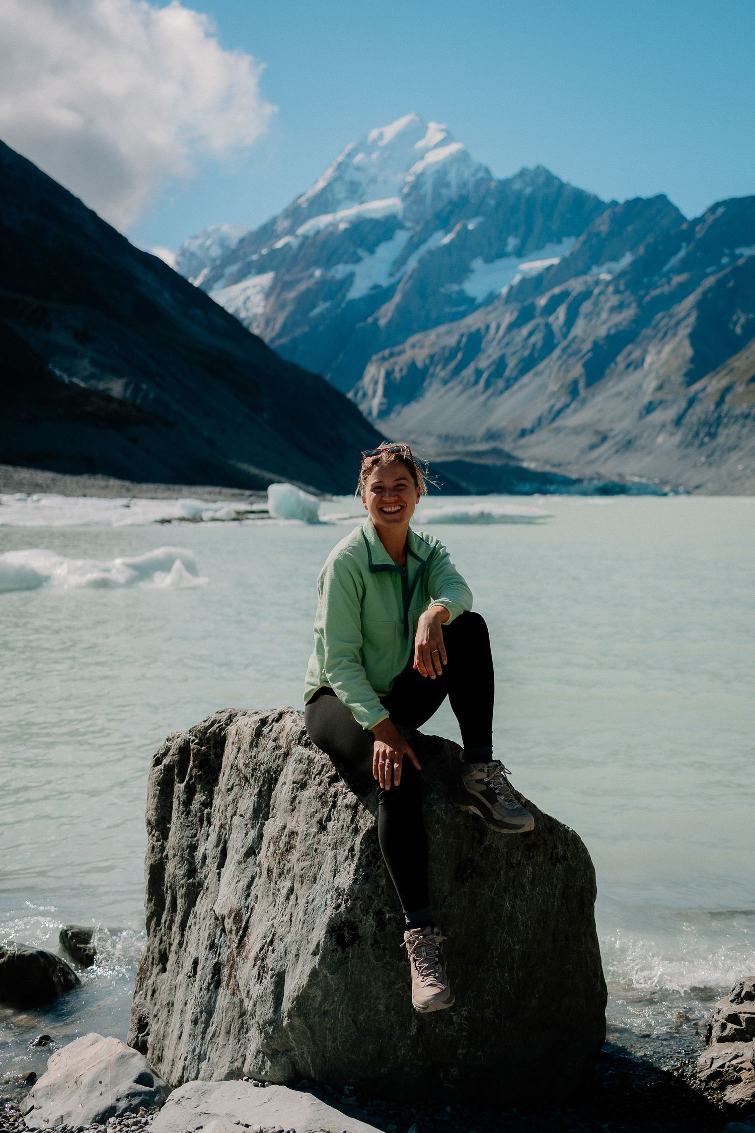 Woman smiling on a rock by a glacial lake with snow-capped Mount Cook in the background, New Zealand.