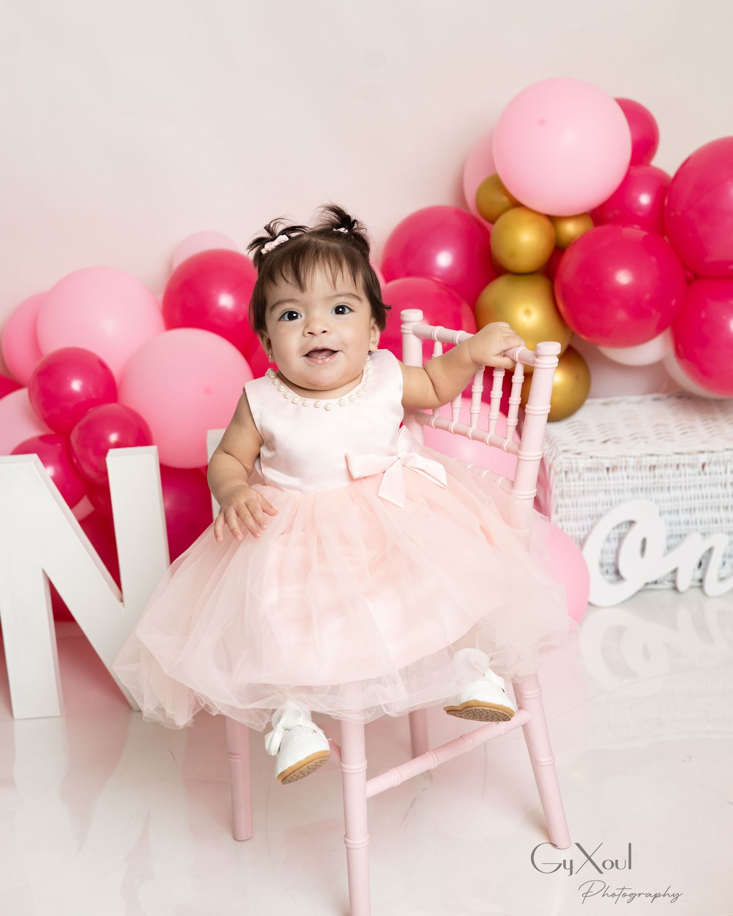 Baby girl in pink tutu dress sits on pink chair at first birthday party with colorful balloon arch backdrop.