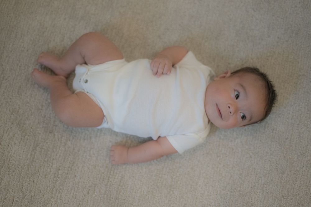 Cute baby in white onesie lying on carpet, looking up with bright eyes and a calm expression.