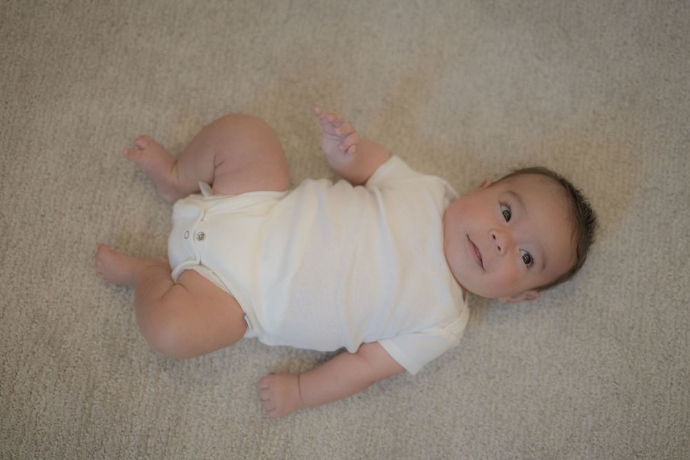 Cute baby in white onesie lying on beige carpet, looking up at camera with bright eyes.