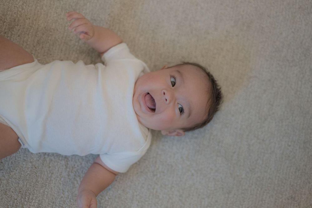 Happy baby in white onesie lying on carpet, smiling with mouth open and arm raised.