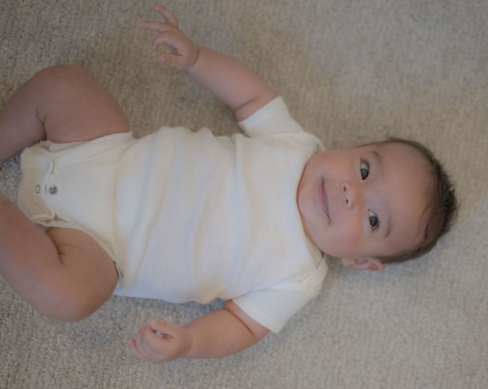 Smiling baby in white onesie lying on carpet with arms raised, looking up at camera.