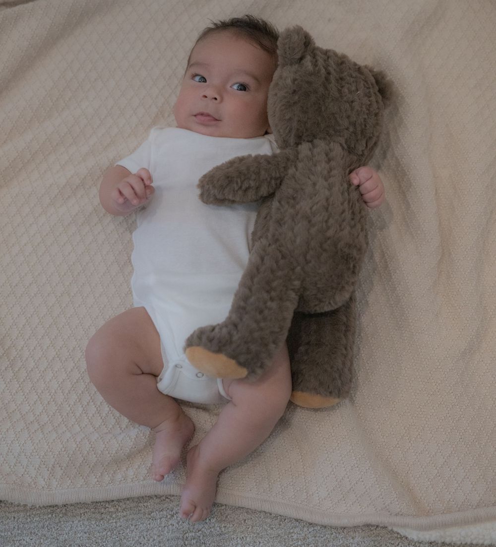 Newborn baby in white onesie lying on cream blanket next to a large brown teddy bear stuffed animal.