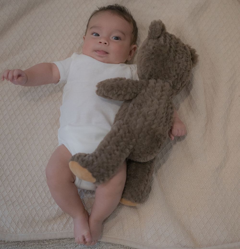 Infant in white onesie lying next to a large brown teddy bear on a white blanket.