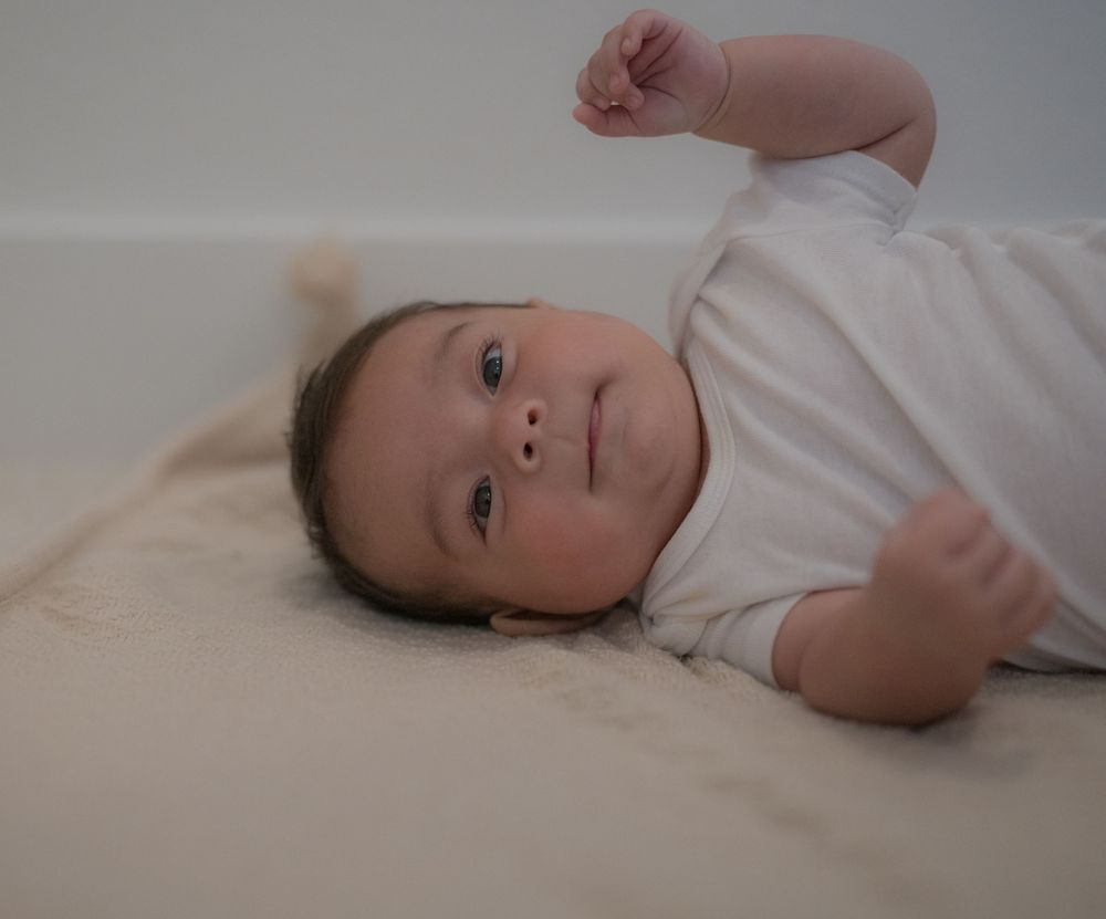 A newborn baby in a white onesie lying on a cream blanket, gazing upward with one fist raised.