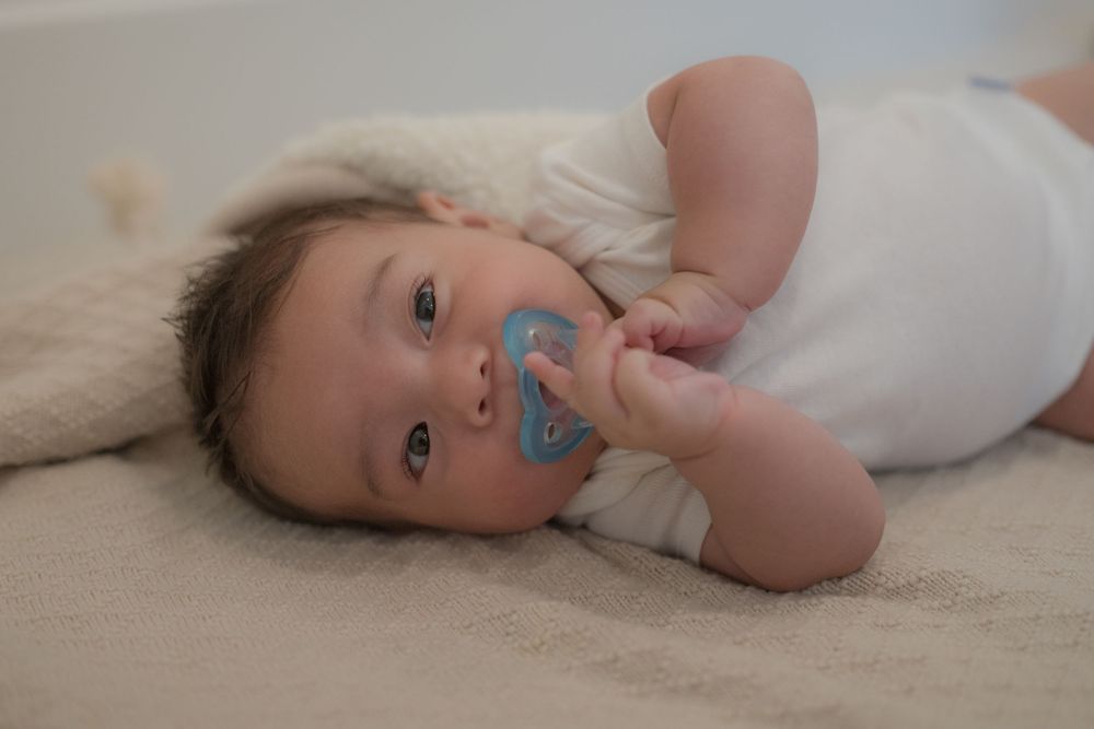 Baby lying on a soft blanket wearing a white onesie, sucking on a blue pacifier, looking up with curious eyes.