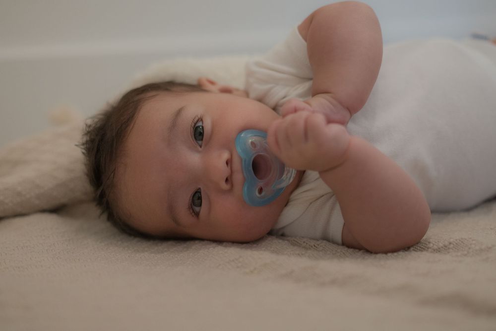 Cute baby lying on a soft blanket with a blue pacifier, looking curiously at the camera.