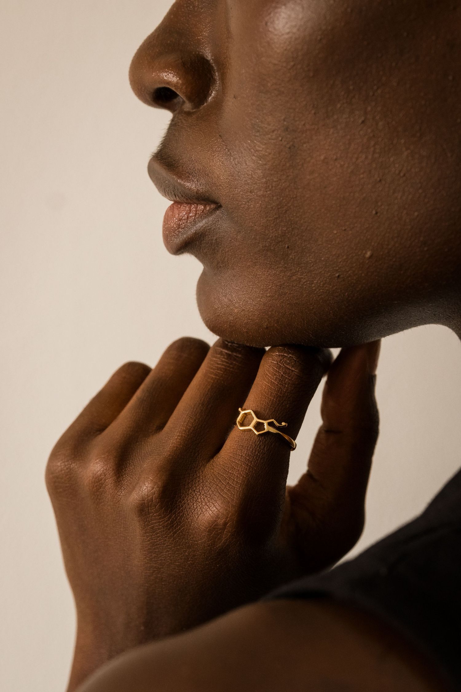 Close-up of a person wearing a gold geometric honeycomb ring, hand resting gracefully near their chin.