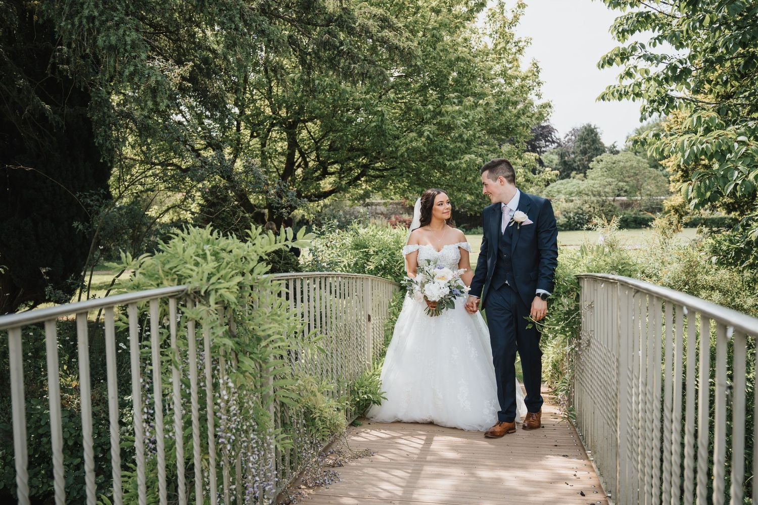 Bride and groom share a romantic moment on a white bridge surrounded by lush green gardens on their wedding day.