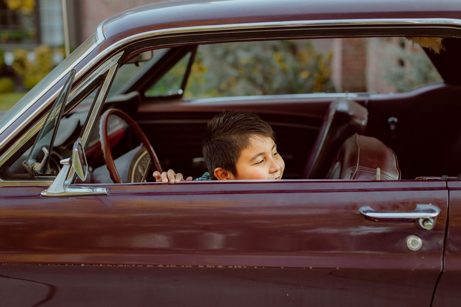 A little boy sits behind the wheel and peeks out the open driver side window in his dad's classic car during a family photo session.