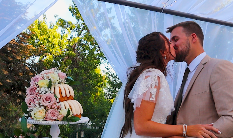 A newlywed couple in wedding attire share a tender kiss while seated outside a building with stained glass windows.