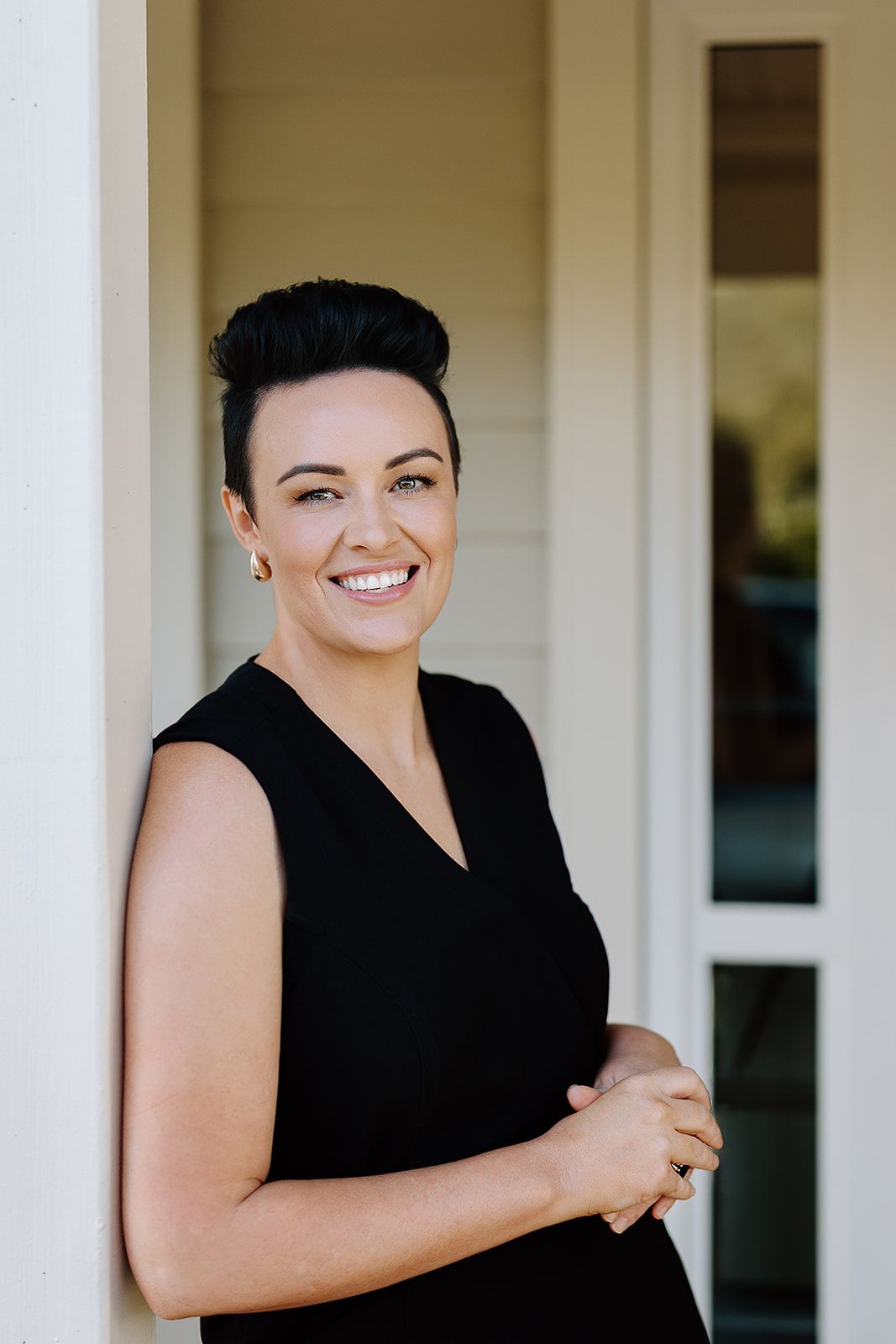 Smiling woman with short dark hair wearing a black sleeveless dress, leaning against a white column outdoors.