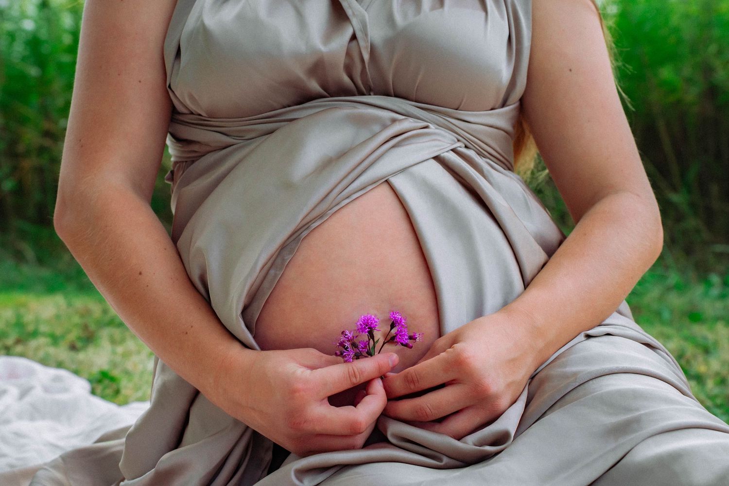 Pregnant woman in gray dress holding purple flowers against her bare belly outdoors.