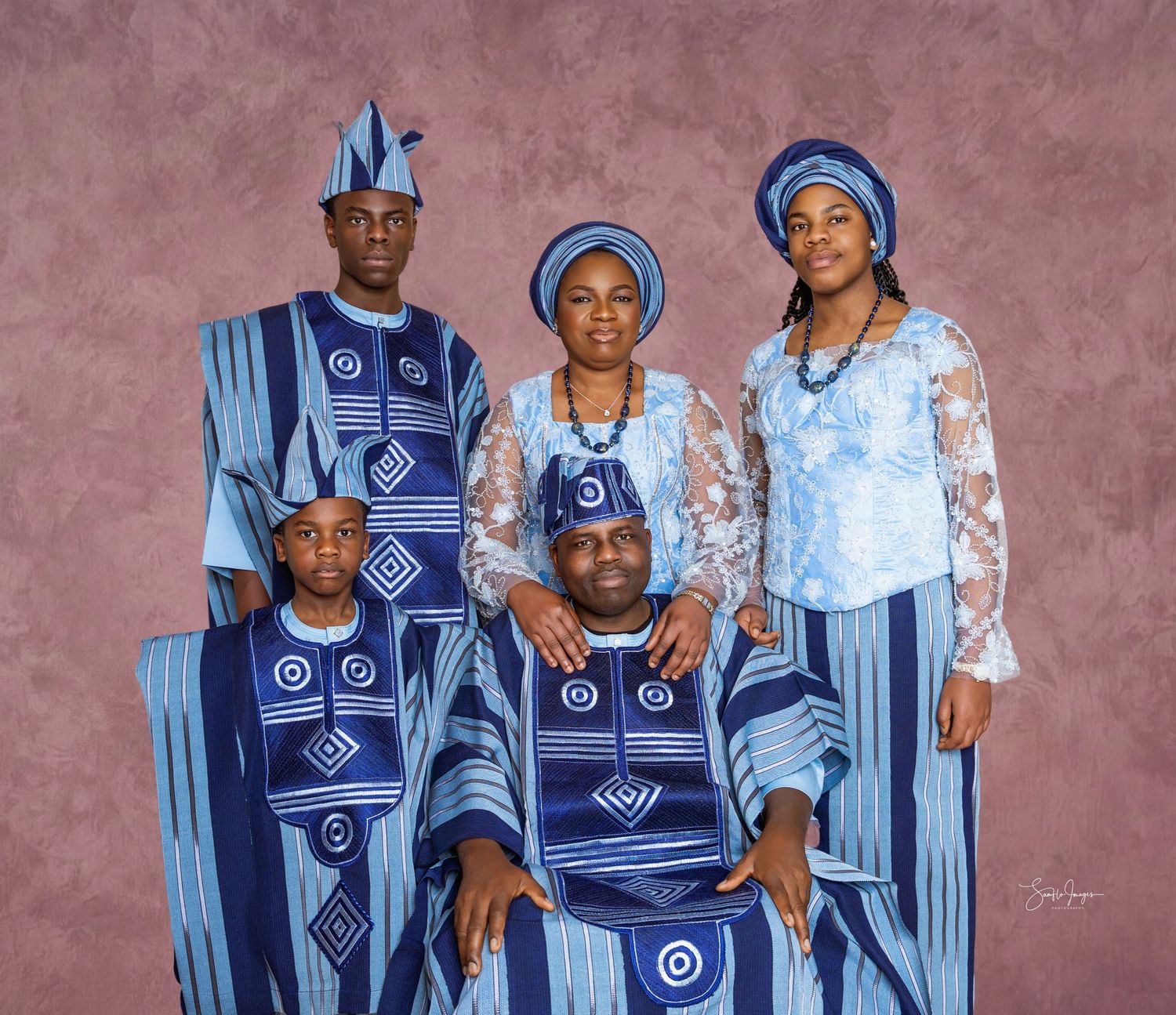 Family of five posing in matching blue and white traditional Yoruba attire with embroidered patterns and coordinated caps against a mauve ba