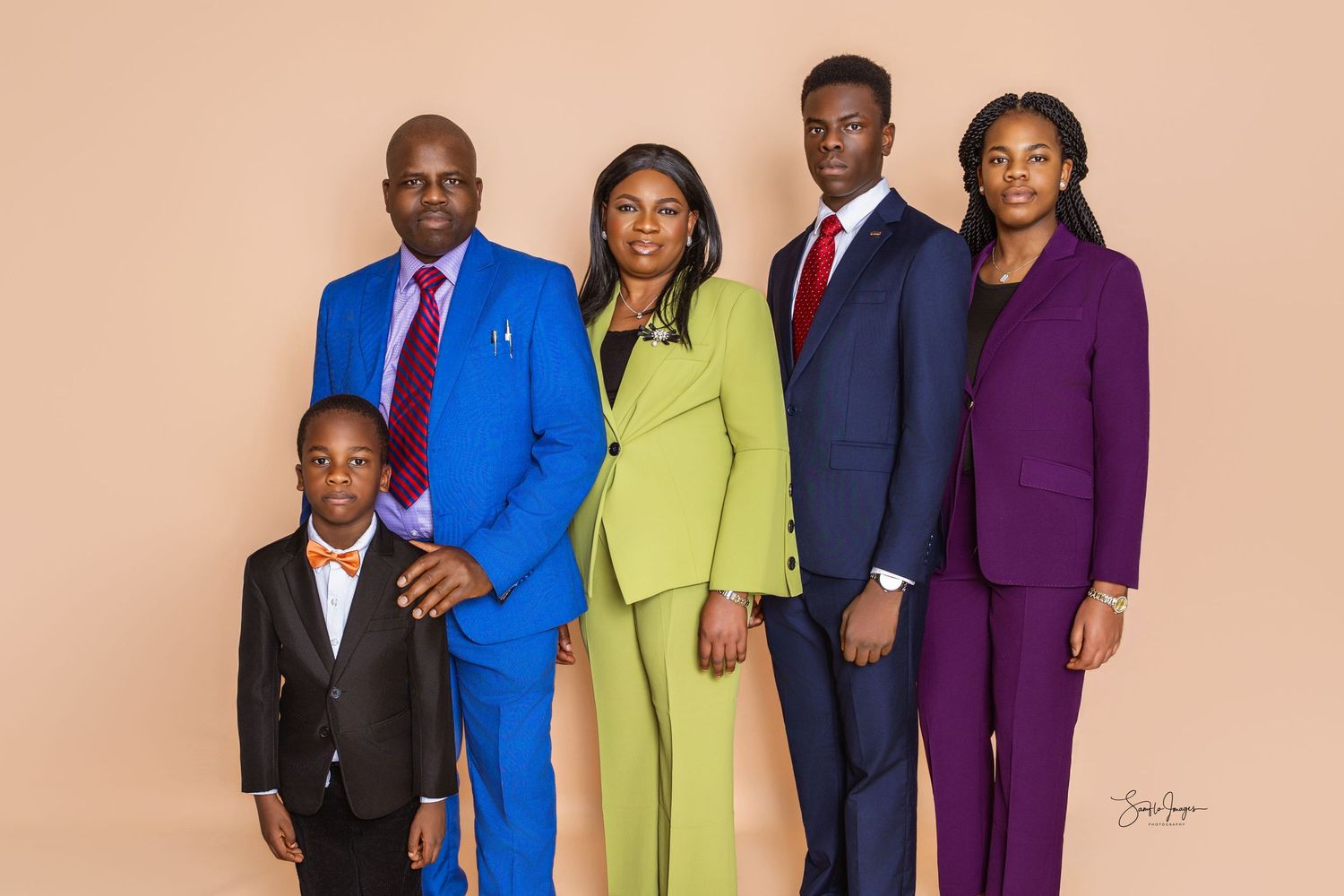 Family of five in coordinated blue, green, navy and purple suits posing together against a peach studio backdrop in a formal portrait.