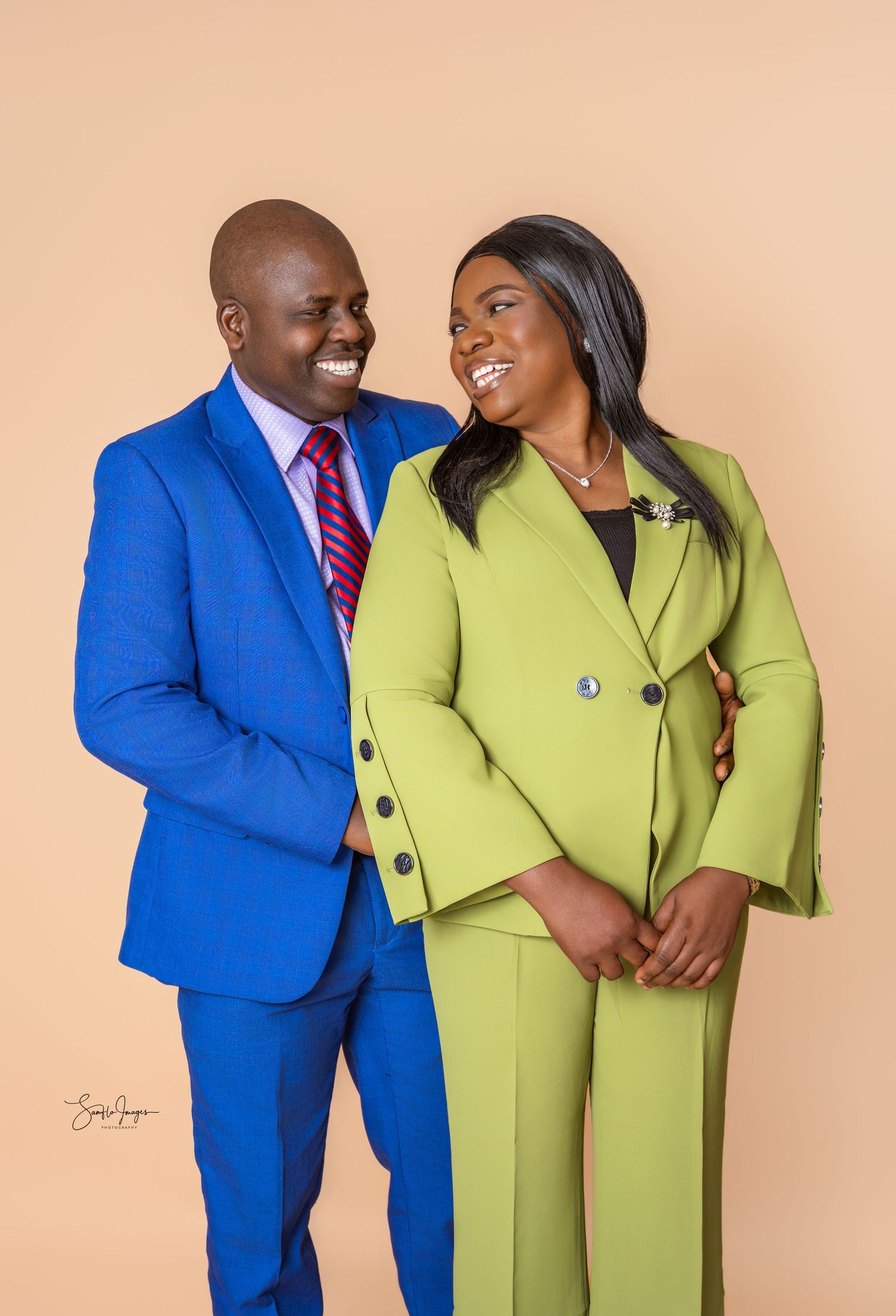 A smiling couple poses together in a studio, the man in a bright blue suit and striped tie, the woman in a lime green pantsuit.