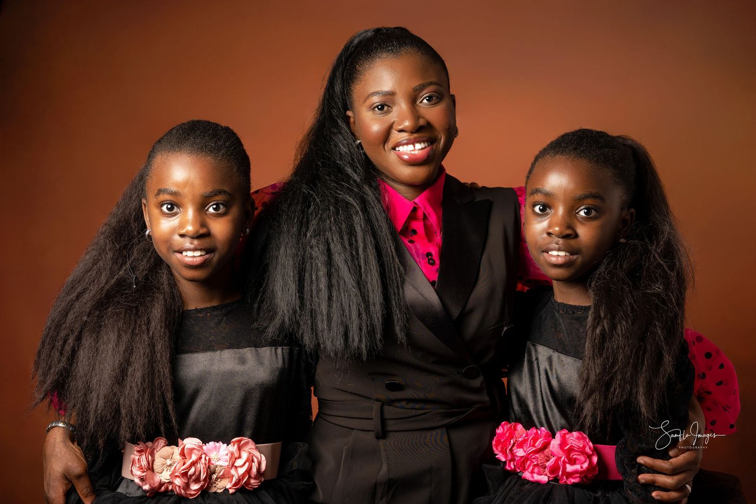A woman and two girls in matching black outfits smile together, holding pink flowers against a warm brown background.