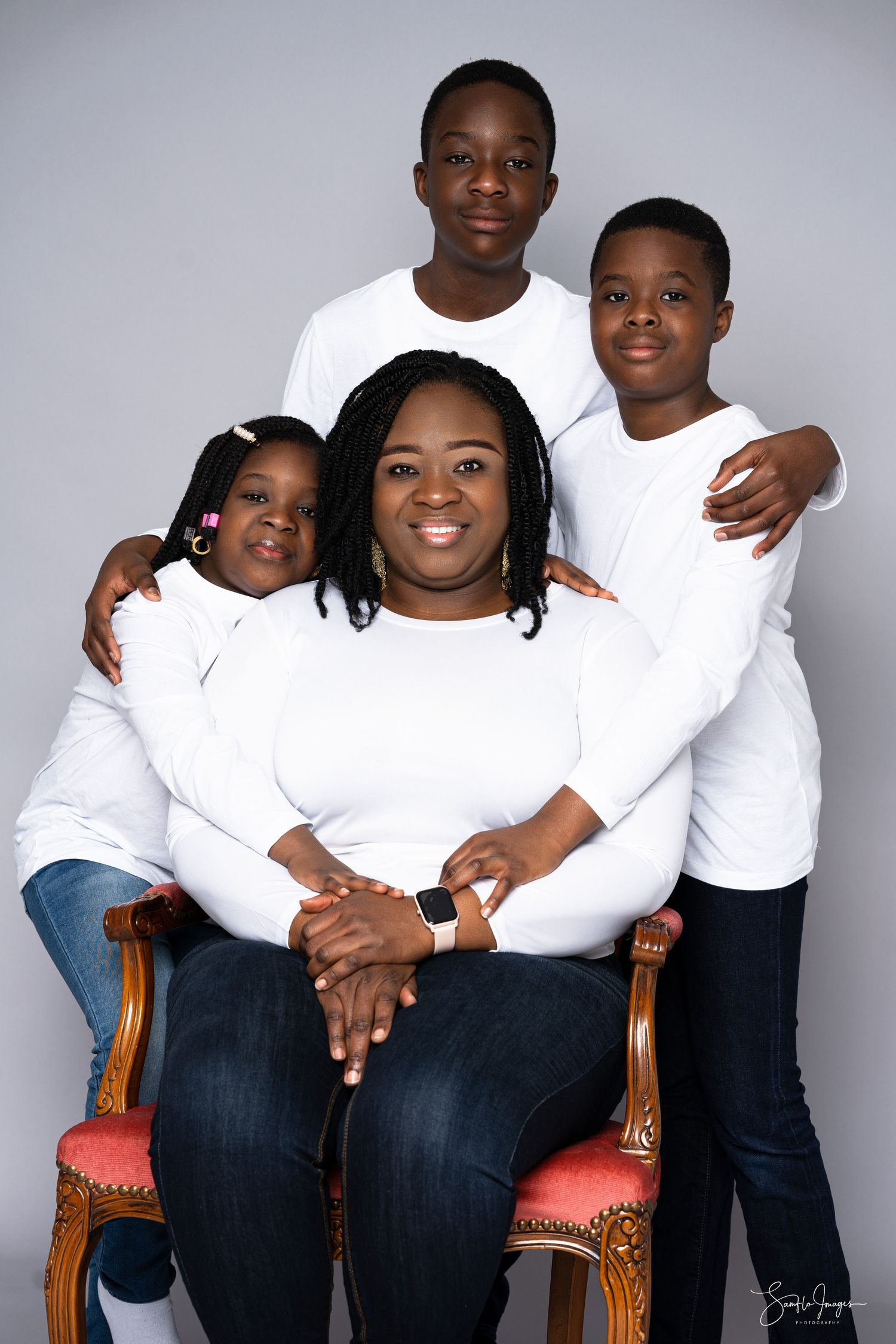Family of four in matching white tops and jeans posing for a studio portrait, mother seated with three children around her.