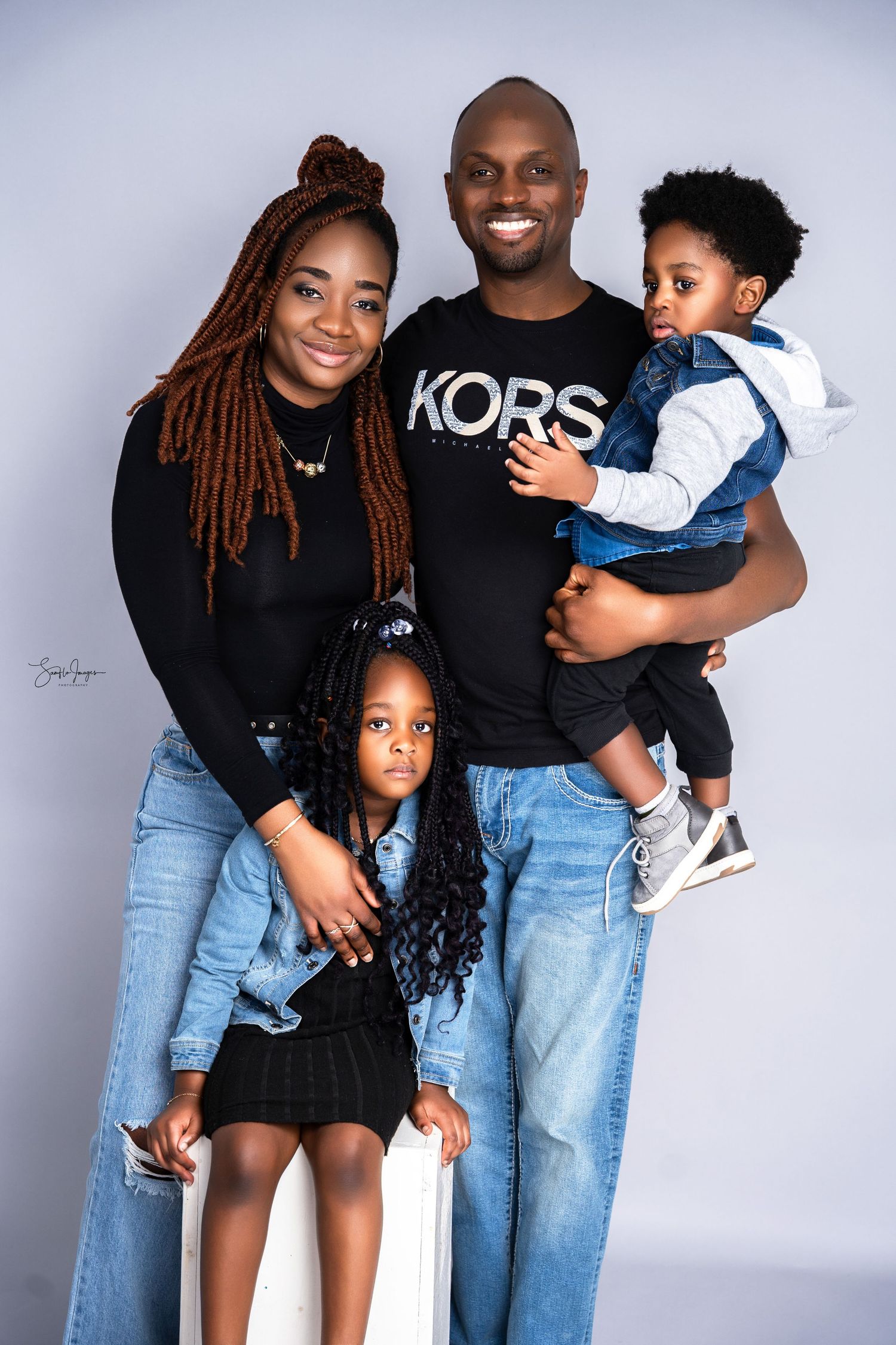 A happy Black family of four poses in a studio, wearing jeans and black tops, with two young children.