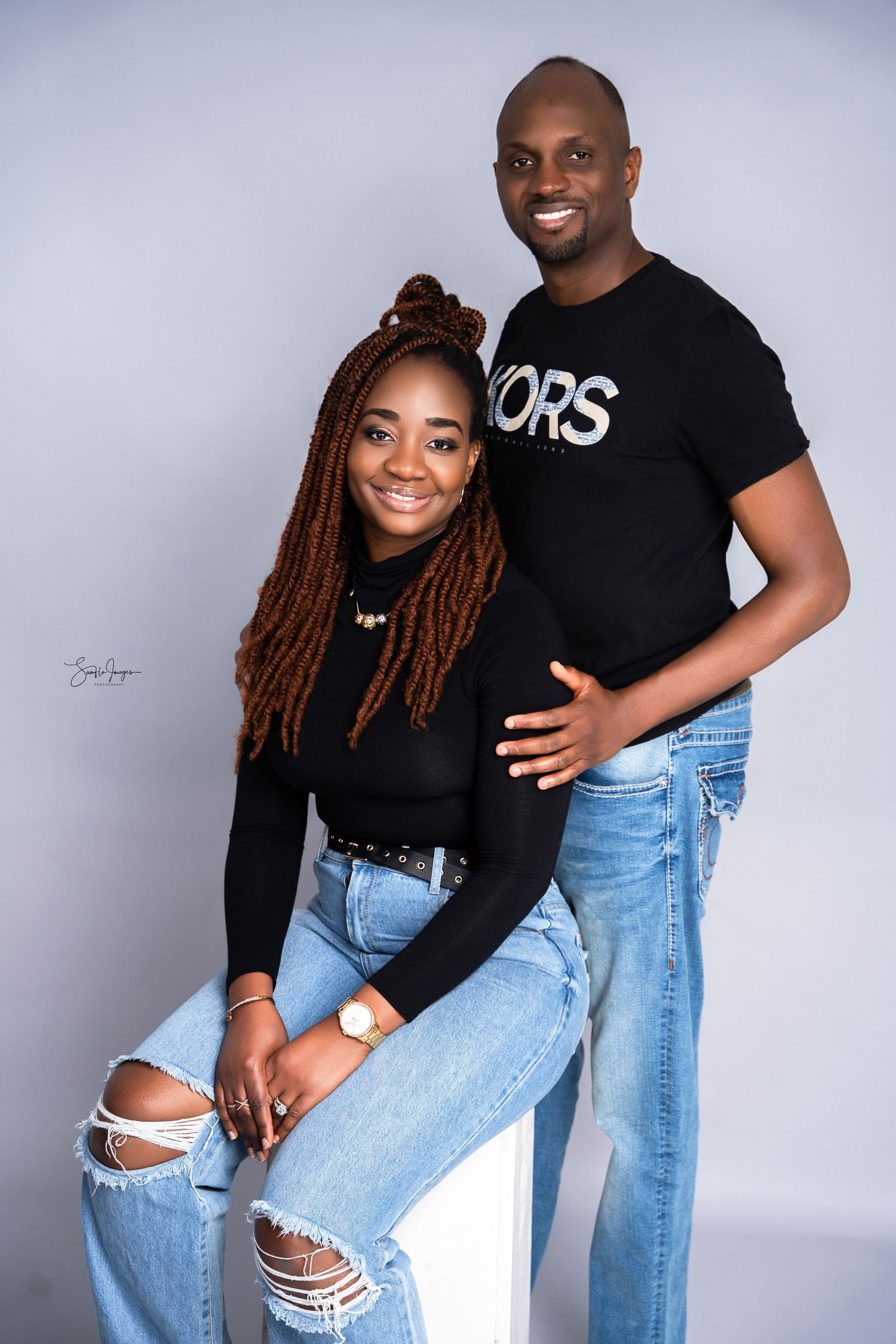A smiling couple in matching black tops and jeans poses together in a professional studio photo shoot.