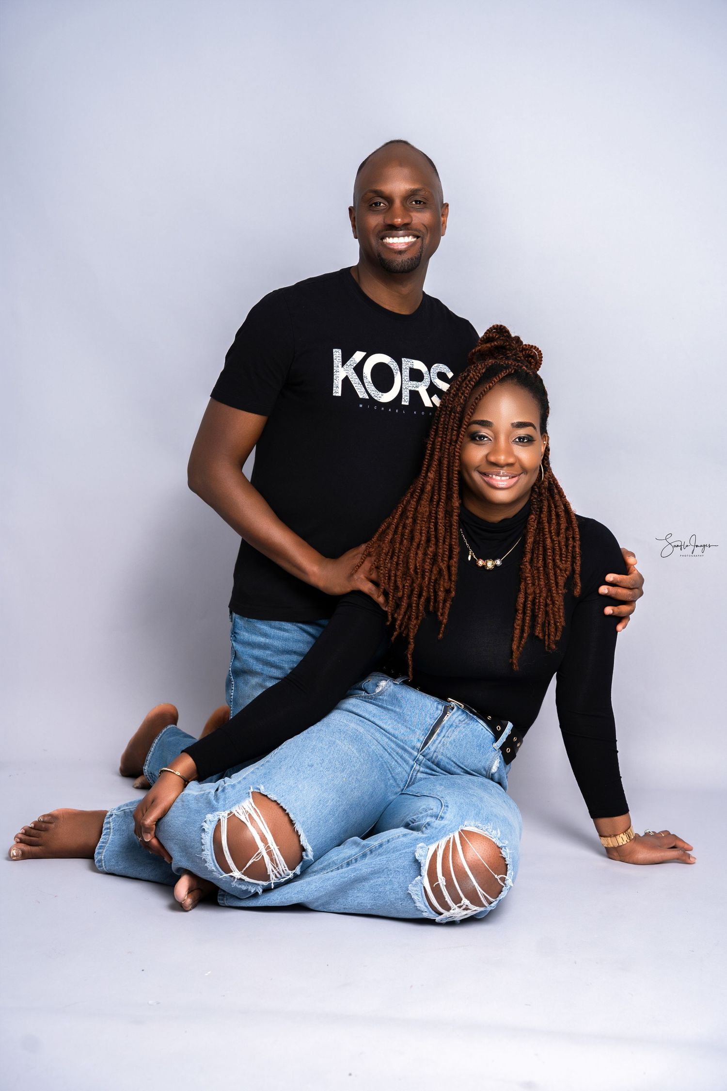 A smiling couple in black tops and ripped jeans pose together against a white studio background.