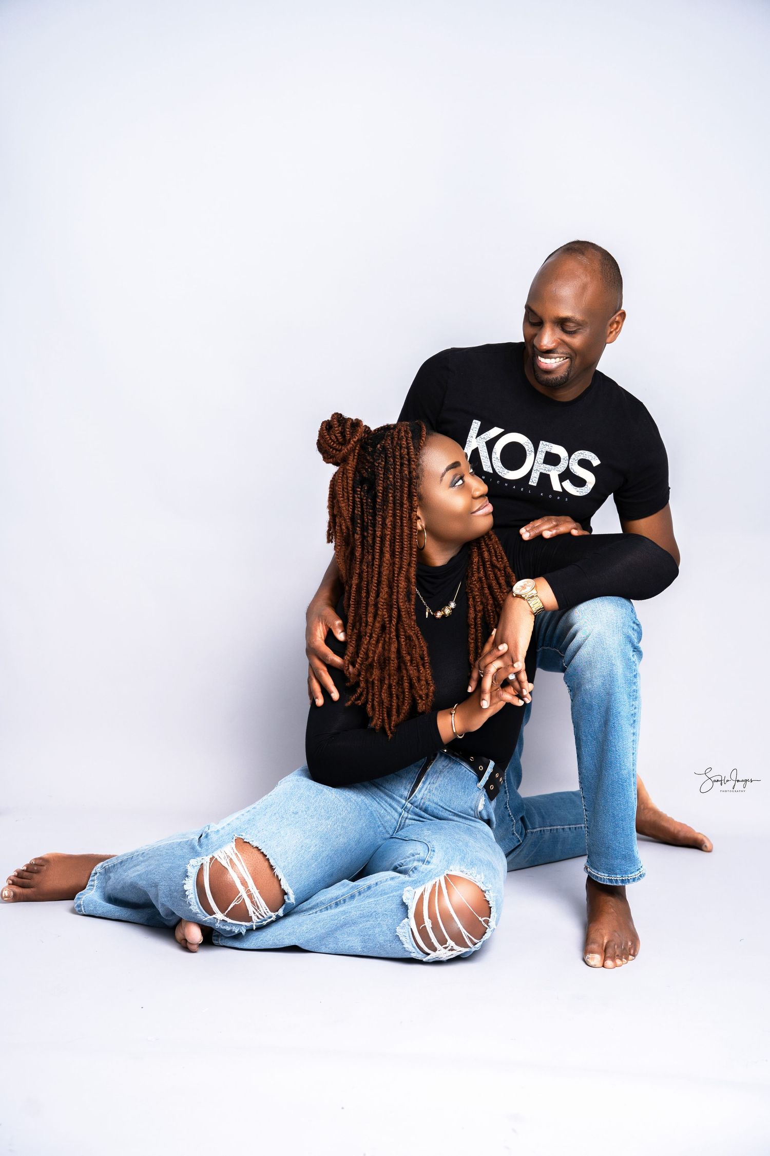 A couple poses together in matching black tops and ripped jeans against a white background in a studio photo shoot.