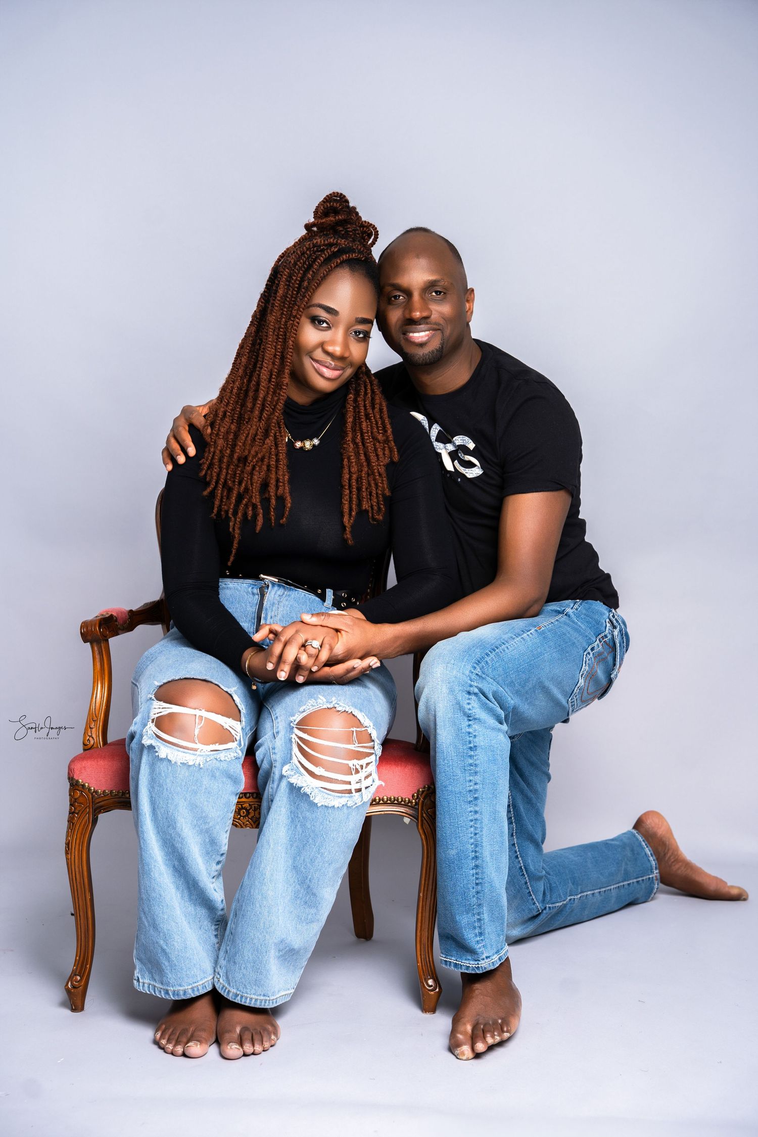 Smiling couple in matching black tops and ripped jeans posing together on a vintage chair in a studio.