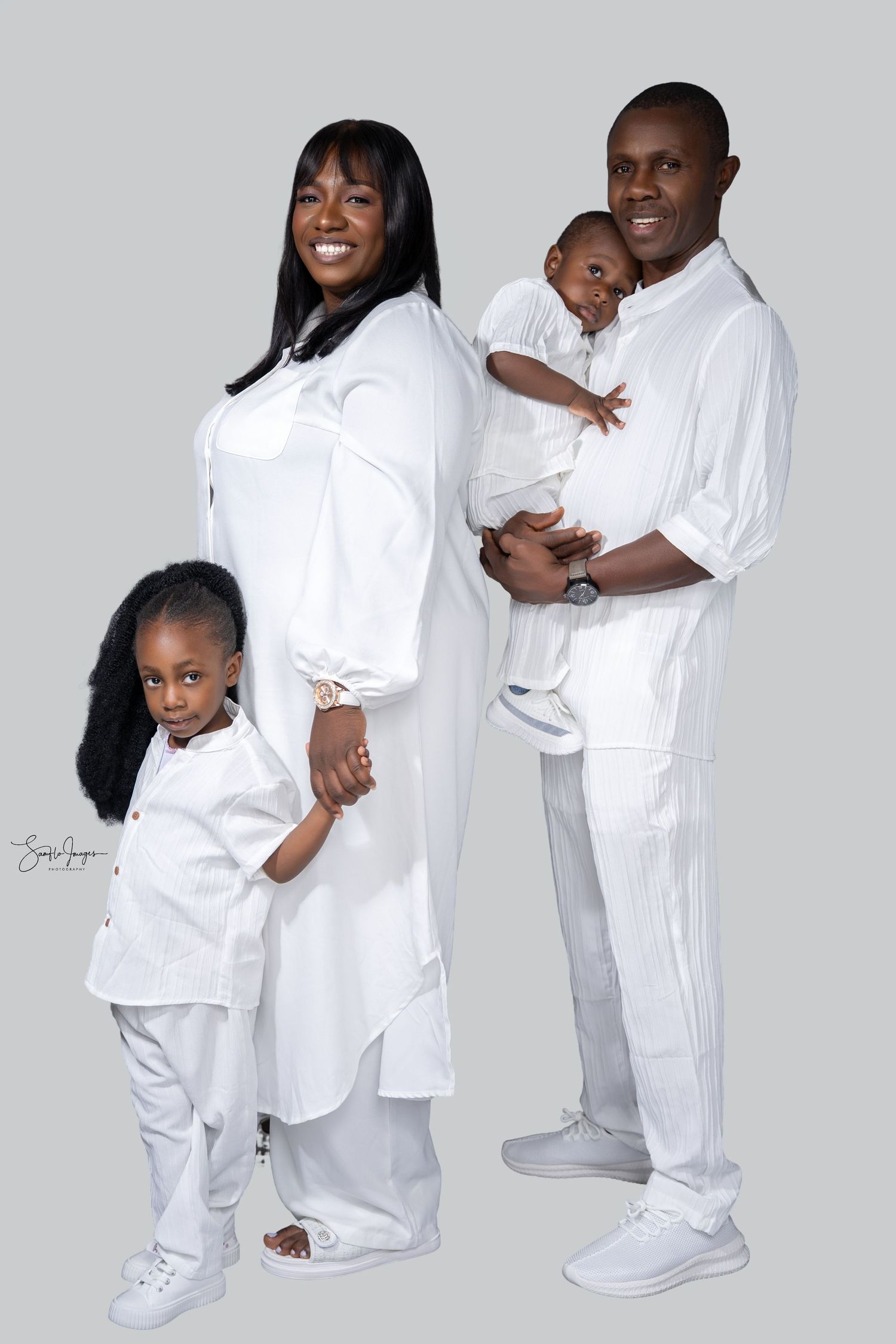 Family of four in matching white outfits posing for a portrait, parents holding children against gray background.